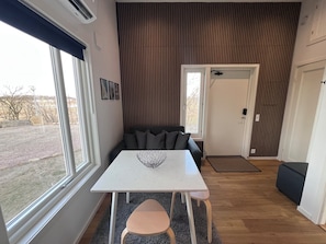 Een moderne woonkamer met een witte tafel, een grijze bank en uitzicht op de omgeving door grote ramen.