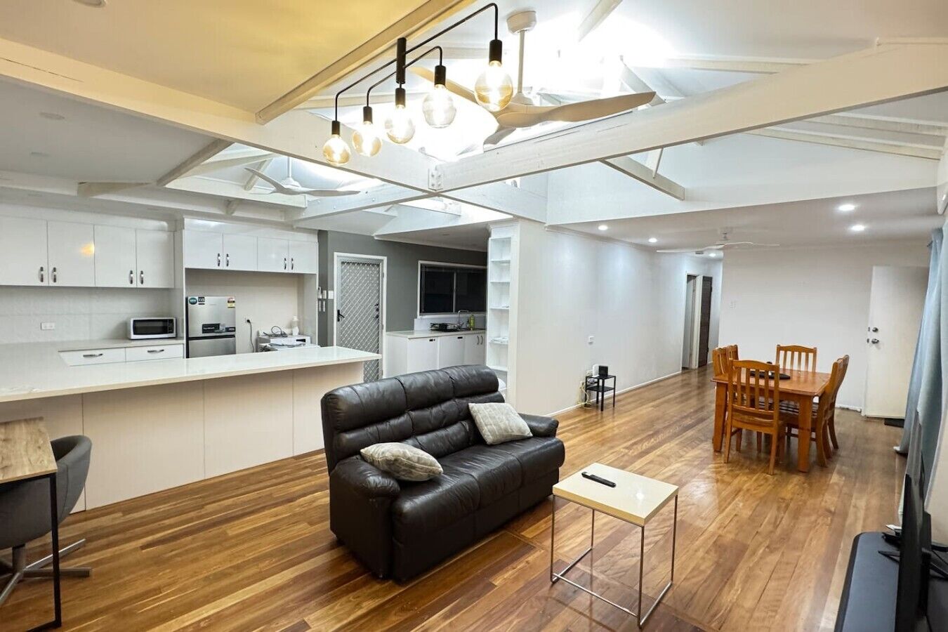A modern kitchen with white cabinets, a dining area with a wooden table and chairs, a leather sofa with cushions, and a small side table.