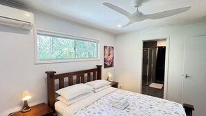 Une chambre à coucher comprenant un lit, des lampes de chevet, un ventilateur de plafond, une fenêtre et une porte donnant sur une salle de bain.