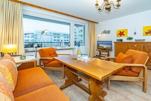 Een kamer met een houten eettafel, oranje stoelen, een bank, een televisie en een raam met uitzicht op gebouwen.