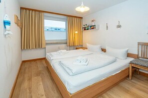 Quarto com duas camas, janela com cortinas, mesa de cabeceira, cadeira e prateleira com objetos decorativos.