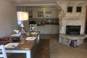 A dining area with a long wooden table set for four, a stone fireplace, and a kitchen in the background. - Holiday apartment with 3 bedrooms (Prerow)