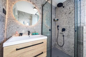 Un bagno moderno con un lavandino bianco, cassetti in legno, uno specchio rotondo e una cabina doccia in vetro.