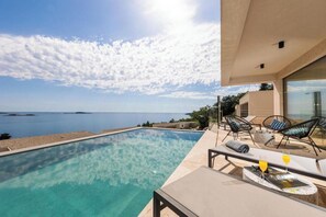 Une piscine offrant une vue dégagée sur la mer, un espace extérieur aménagé pour se détendre et une façade de bâtiment au design moderne.