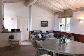 Een gezellige kamer met een ronde tafel, rieten stoelen, een bank met kussens, een klein krukje, een witte deur en een raam met uitzicht.