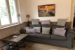 Een woonkamer met een grijze bank, een glazen salontafel en een schilderij van een zonsondergang boven de zee.