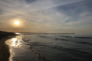 A beach with a pier extending into the water, waves gently lapping the shore, and the sun setting in the background. - Prerow || beach house (Prerow)
