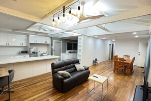 Uma cozinha moderna com armários brancos, uma área de jantar com mesa e cadeiras de madeira, um sofá de couro com almofadas e uma pequena mesa lateral.