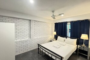 Quarto de hotel com parede de tijolos, cama com roupa branca, duas mesas de cabeceira com luminárias e um ventilador de teto.