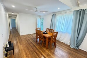 Comedor con una mesa y sillas de madera, un ventilador de techo y vista a través de una ventana con cortinas.