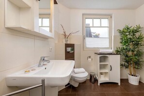 Un baño moderno con un lavabo, inodoro y mueble blancos, una ventana y una planta en maceta.