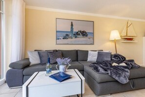 Een woonkamer met een grijze bank, een witte salontafel, een schilderij van een vuurtoren, een lamp en een klein model zeilboot.