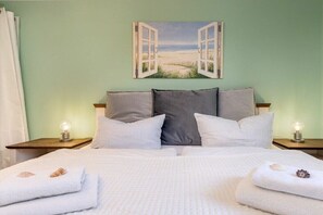 Een slaapkamer met een bed, kussens, een schilderij van een open raam en bedlampjes.