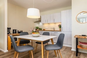 Une cuisine compacte avec un coin repas, des meubles blancs, un luminaire suspendu et une petite table avec un panier.
