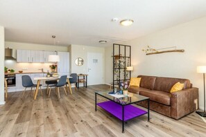 Een moderne woonkamer met een glazen salontafel, een bruine leren bank en een eethoek met een witte tafel en stoelen.