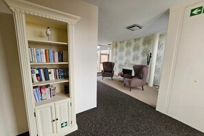 Een kamer met een boekenkast, twee fauteuils, een open haard en een vloerbedekking.