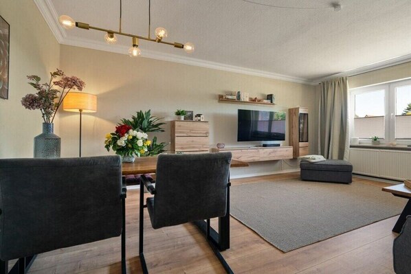 Una sala de estar moderna con un televisor de pantalla plana, una mesa de comedor con sillas, un sofá, una lámpara y un jarrón decorativo con flores.
