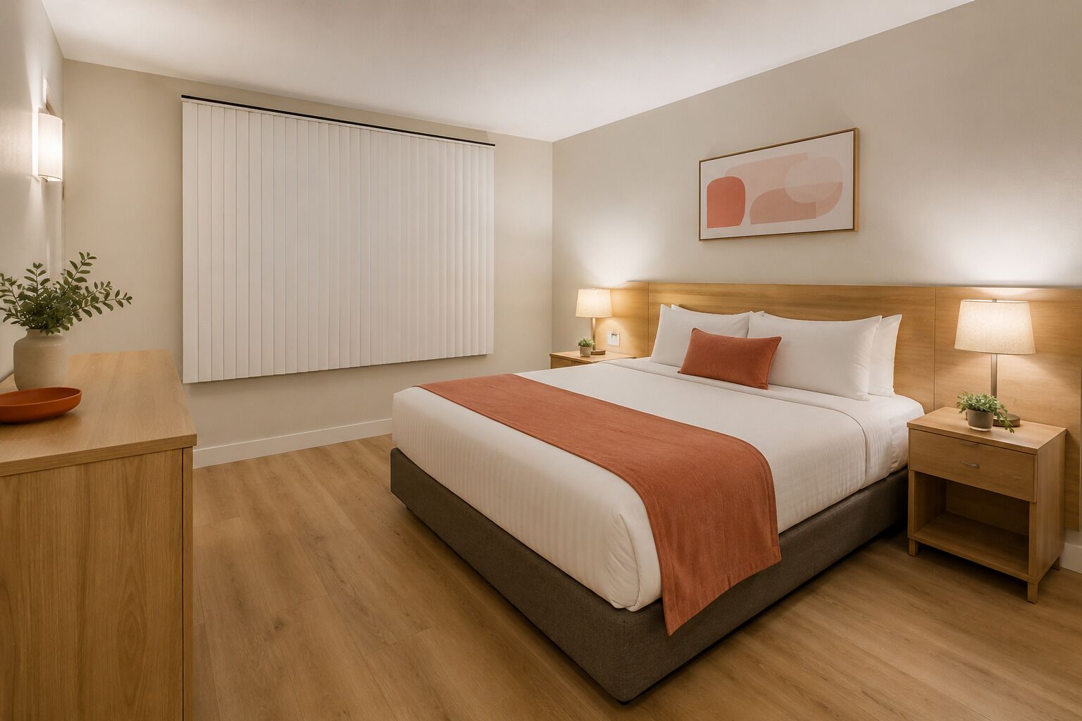 A hotel room with a bed, bedside tables, a window with blinds, a potted plant, and a wall-mounted artwork.