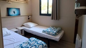 Una habitación pequeña y bien iluminada con dos camas individuales, una ventana con cortinas, un estante en la pared y una lámpara colgante.