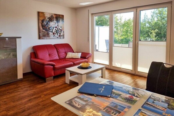 A living room with a red sofa, a small table with a book, and a large window with a view of trees. - Holiday apartment with 1 bedroom (Sassnitz)