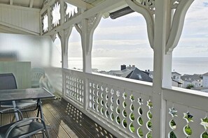 Ett takförsett utomhusområde med ett bord och stolar, ett vitt räcke med dekorativa utskärningar och utsikt över havet och byggnaderna i bakgrunden.
