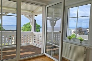 Un balcon avec vue sur la mer, une balustrade blanche et une fenêtre ornée d’une plante.