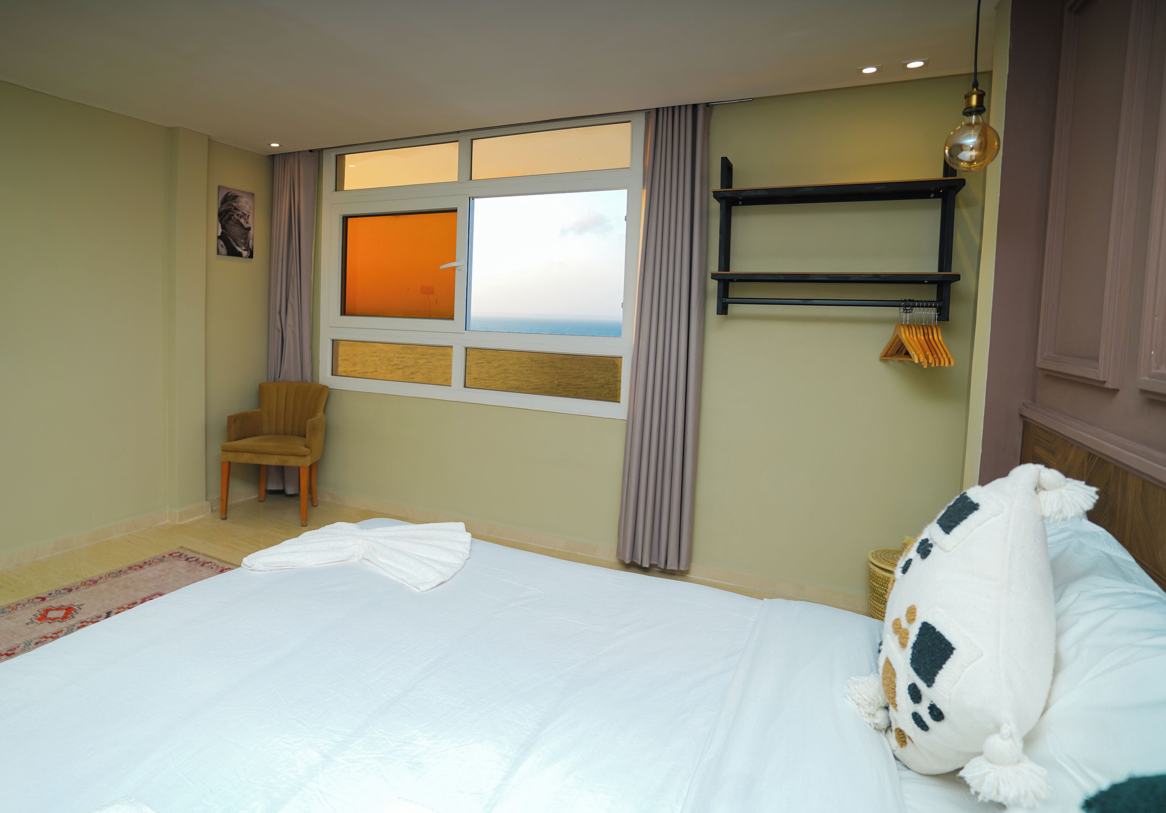 A hotel room with a large bed, a chair, a window with curtains, a shelf, and a hanging light fixture.