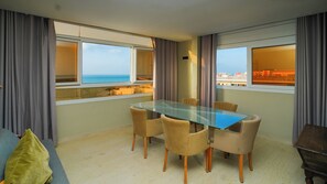 Habitación de hotel con una mesa de vidrio, cuatro sillas, un sofá y ventanas grandes con cortinas.