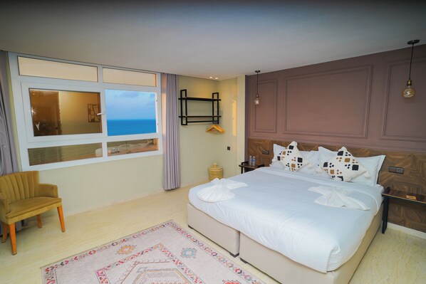Habitación de hotel con una cama grande, una silla, una mesita de noche, una lámpara colgante y vistas al mar a través de una ventana.