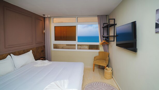 Une chambre d’hôtel avec un grand lit, un téléviseur fixé au mur, une fenêtre donnant sur la mer et un fauteuil.