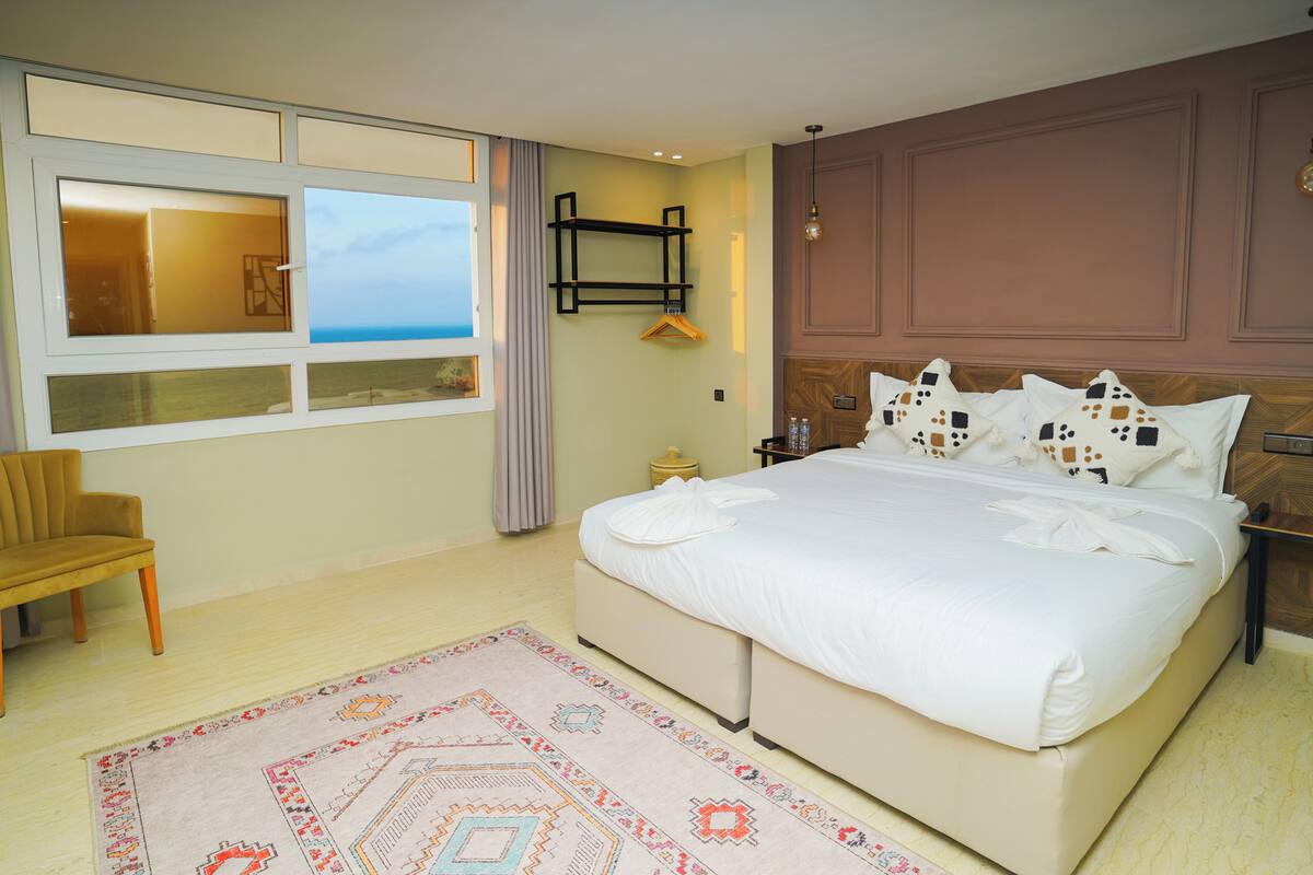A hotel room with a large bed, a chair, a bedside table, a nightstand, a window with a view, and a wall-mounted shelf.