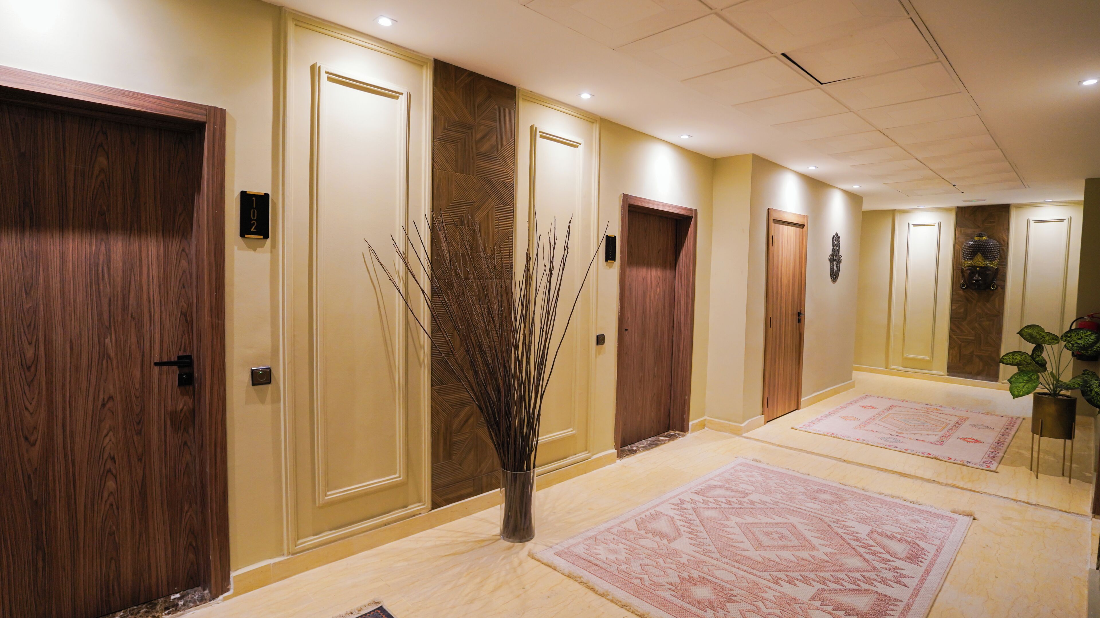 A hotel hallway with wooden doors, a vase with branches, and patterned rugs.