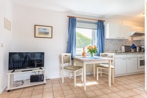 Une cuisine compacte avec des meubles blancs, une table à manger avec des chaises, un téléviseur posé sur un meuble et une fenêtre agrémentée de rideaux bleus.