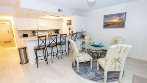 Cocina compacta con armarios blancos, una barra con taburetes, una mesa de comedor con sillas y un cuadro en la pared.