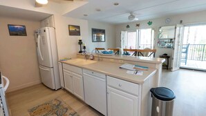Una cocina con refrigerador, lavavajillas y un cubo de basura. Hay una ventana con persianas, un ventilador de techo y una puerta que da a otra habitación.