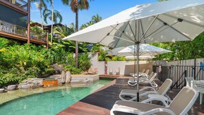 Un área de piscina con sillones de descanso y sombrillas, rodeada de exuberante follaje y una terraza de madera.