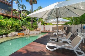 Ein Poolbereich mit Liegestühlen und Sonnenschirmen, umgeben von üppiger Vegetation und einer Holzterrasse.