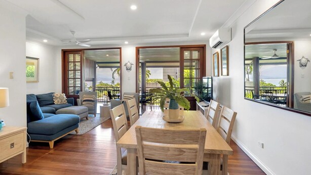 Una sala de estar moderna con una mesa de comedor, sofá, sillas, una mesita auxiliar con lámpara y ventanas amplias con vistas.