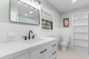 Un bagno con un lavandino bianco, un rubinetto nero e uno specchio grande. Si vede anche un water e una finestra con persiane.