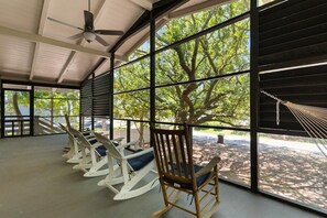 A covered outdoor seating area with rocking chairs and a hammock, large windows with a view of trees and a body of water. - Family Beach Escape 5BR Fenced Yard, Walk to Ocean (Pawleys Island)