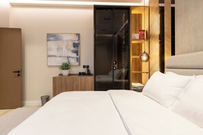 Quarto de hotel moderno com cama, mesa de cabeceira com planta, pintura abstrata na parede, guarda-roupa e porta de vidro para o banheiro.