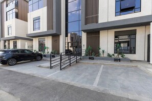 Um edifício moderno com um carro preto estacionado na frente, uma escadaria que leva à entrada e uma pequena área de jardim com plantas.