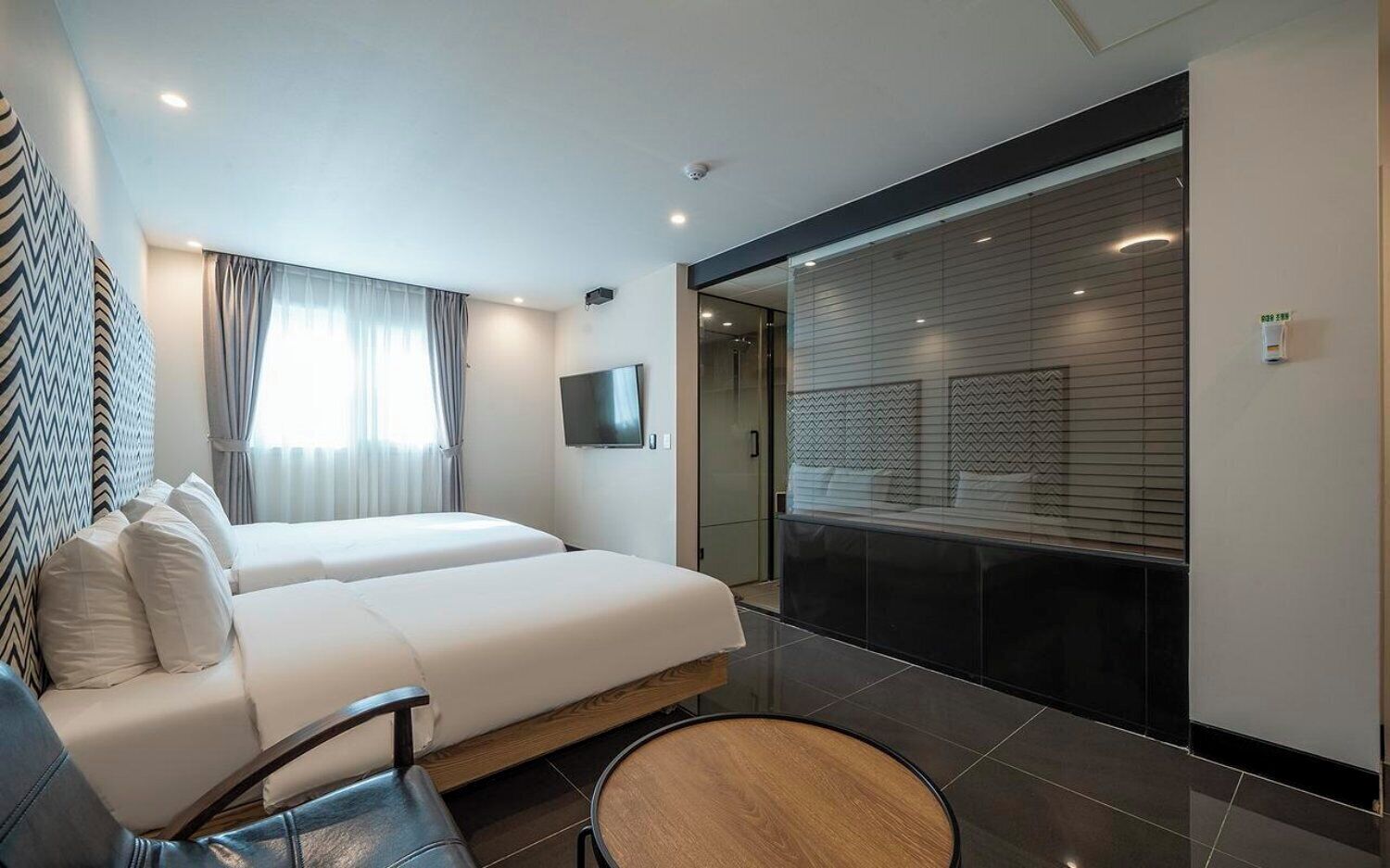 A modern hotel room with a large bed, a TV, a seating area, and a view of the city through the window.