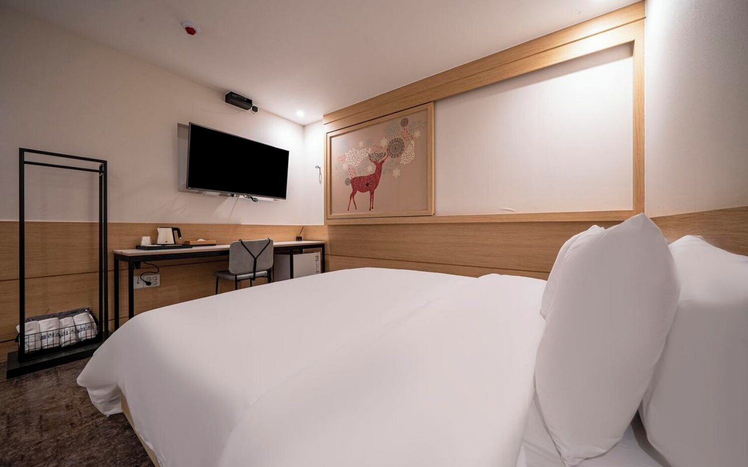A hotel room with a large bed, a desk with a chair, a flat-screen TV mounted on the wall, and a framed artwork above the bed.