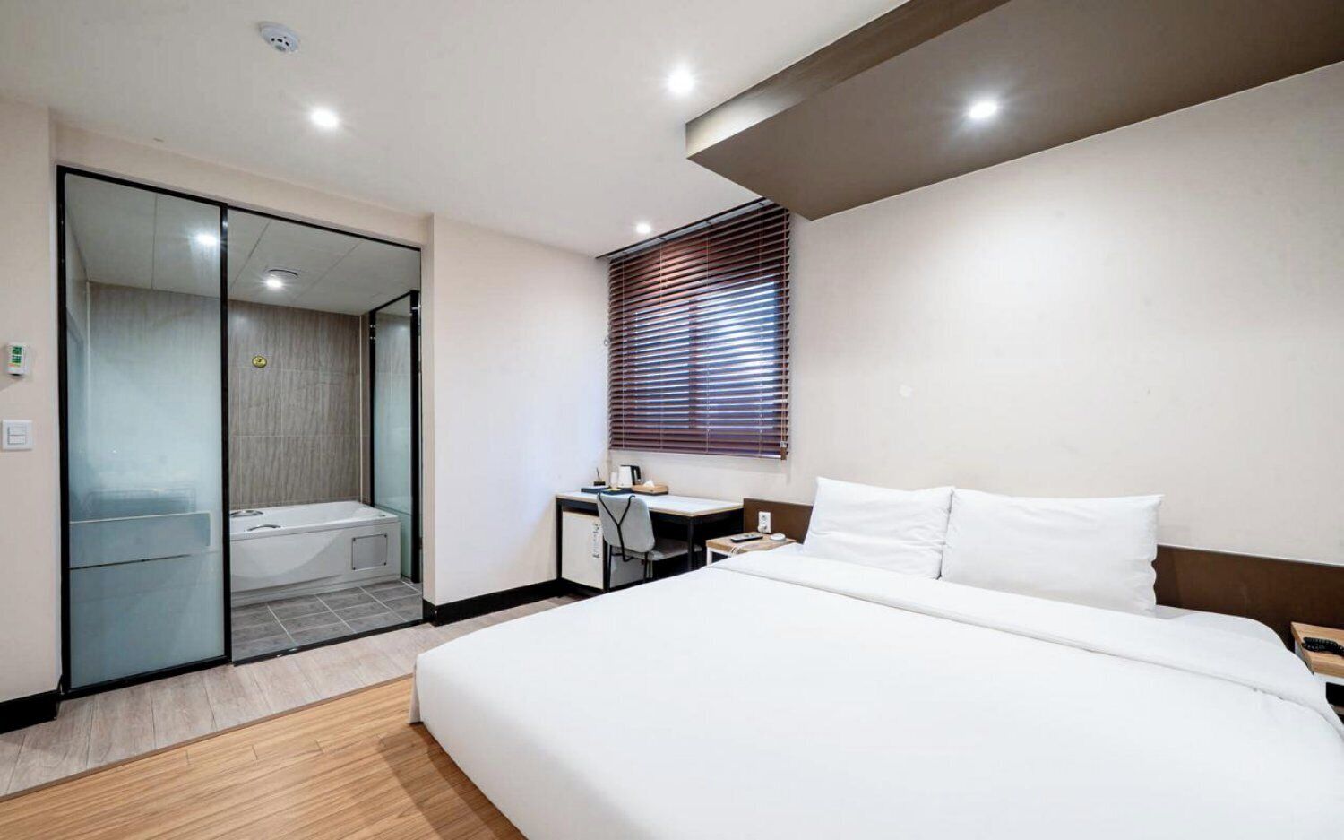 A modern hotel room with a large bed, a desk, a shower, and a window with blinds.