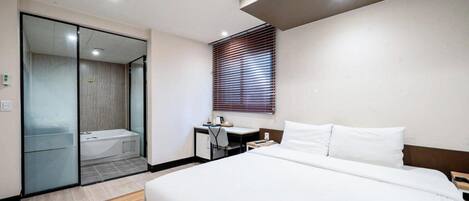 A modern hotel room with a large bed, a desk, a shower, and a window with blinds.