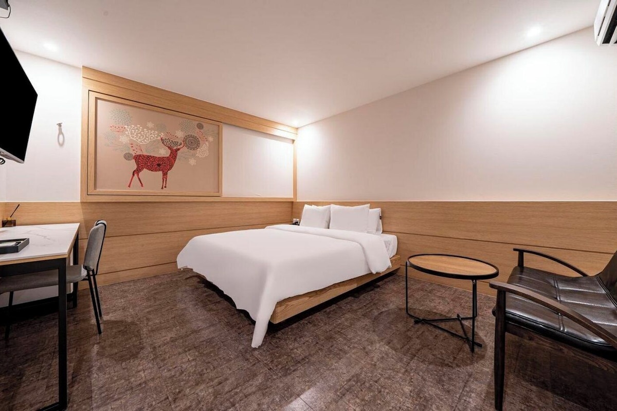 Habitación de hotel con cama, escritorio, silla, mesita y televisor montado en la pared.