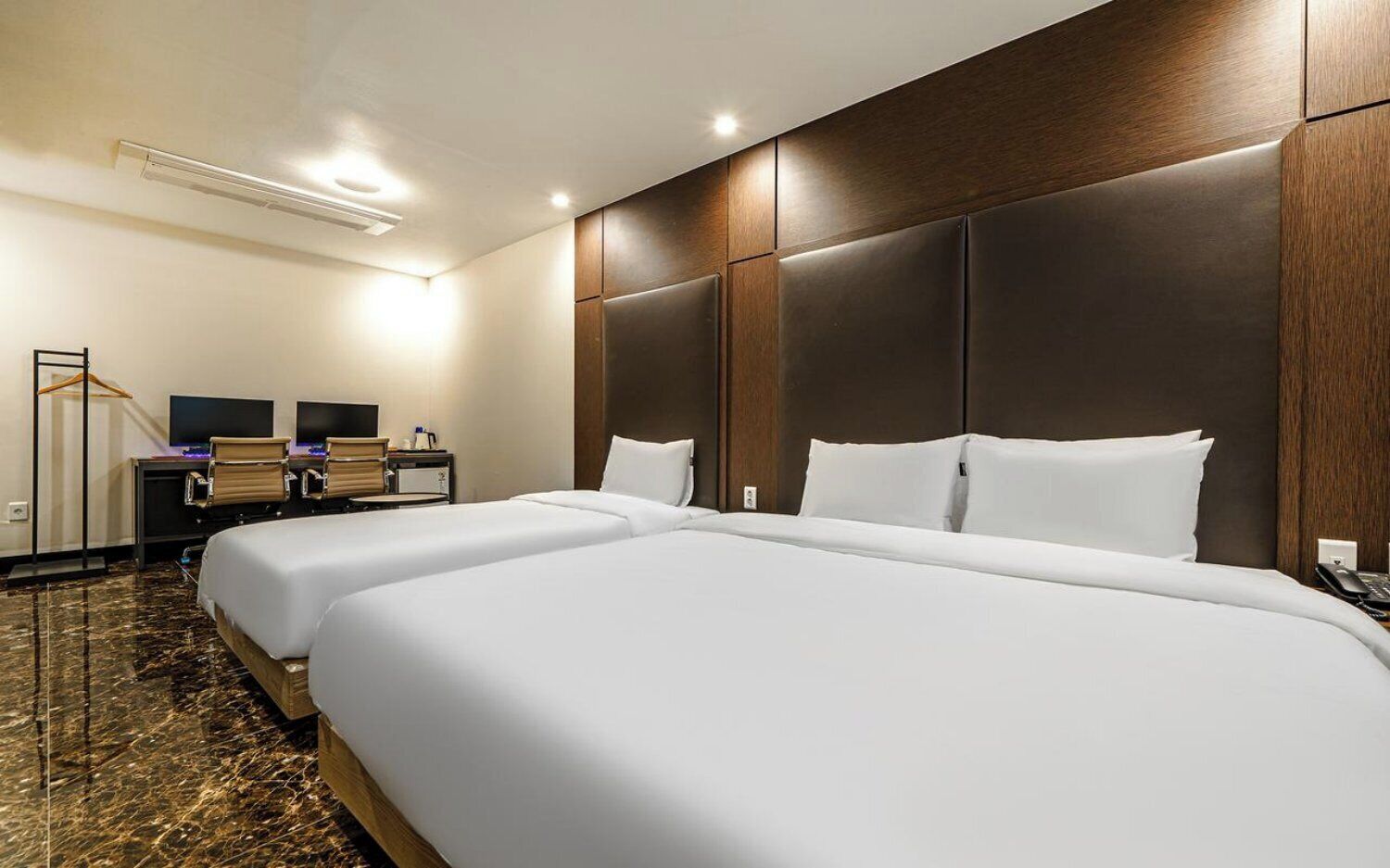 A hotel room with two beds, a desk with two monitors, a coat rack, and a telephone.