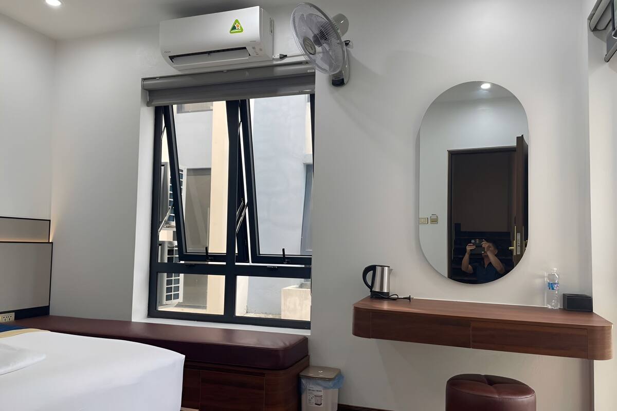 A hotel room with a bed, a window, a mirror, a bedside table with a kettle, and a small stool.