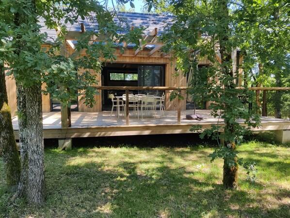 A wooden cabin with a covered patio, a dining area with chairs and a table, and a large window.